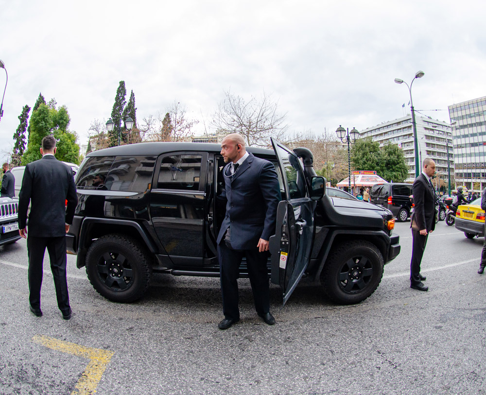 A black car parked with an open door as a man wearing a black suit, black tie and an ear piece is leaving the vehicle. Another man wearing the same clothes is stood at the front of the car facing away from it and another man is stood at the back of the car.