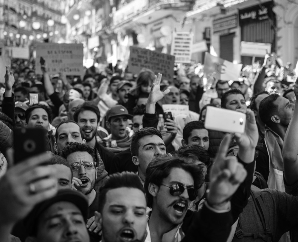 A black and white image shows a large group of people taking park in a protest as they are all stood together holding signs, phones and shouting and pont fingers.