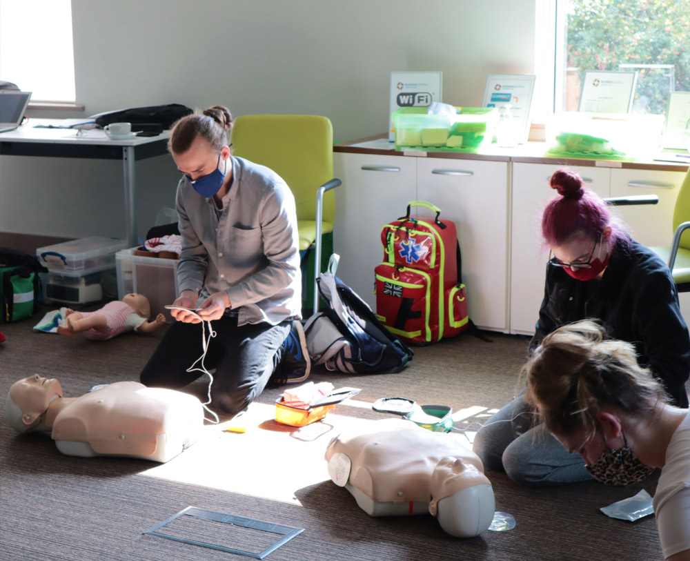 A group are learning how to do CPR on a dummy in the first aid training. A man can be seen getting shock pads to apply to the dummy and the dummies lay on the floor in front of them.