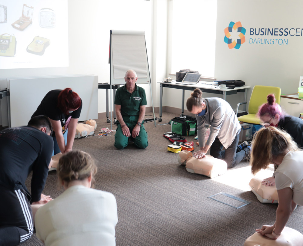 A first aid medic is teach a group how to do CPR on a dummy, The medic is sat in the centre as the students are set next to him continuing in a circle shape. The leaners are demonstrating what they have learnt through CPR on a dummy.