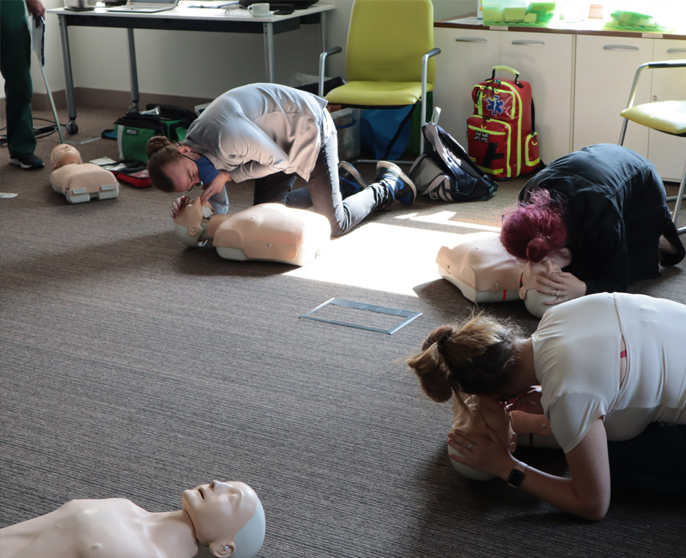 A male learner in a first aid course is seen leaning over the CPR dummy to heck their air waves are hoping by living the chin with their fingers and places their ears near the dummies face to hear them. Two female learners are doing the same task to the risk of the male learner.