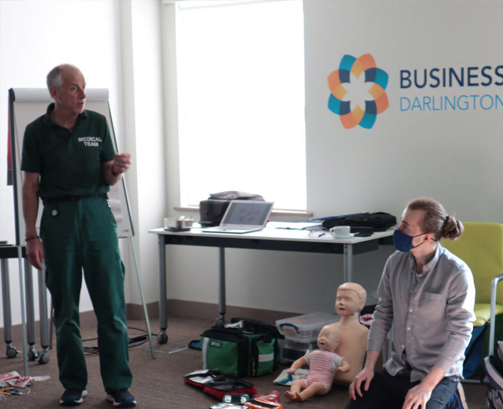 A first aid tutor is wearing a medical team uniform at a first aid training course, He is teach the group how to do CPR including on children. A male learner is looking up listening to him with two child CPR dummies propped up next to him.
