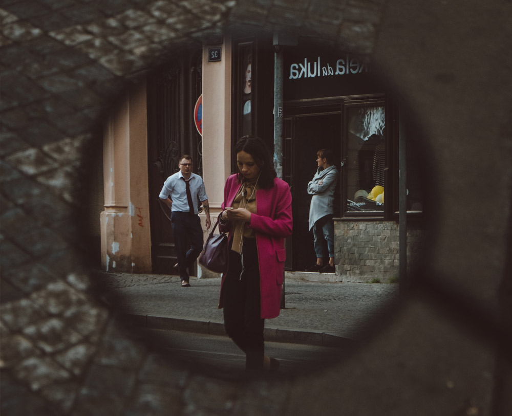 On a street a women can be seen through a zoomed in car wing mirror which looks like a magnifying glass. She is wearing a pink mid length coat as she looks at her phone crossing the street. From this point of view it looks like she is being spied on.