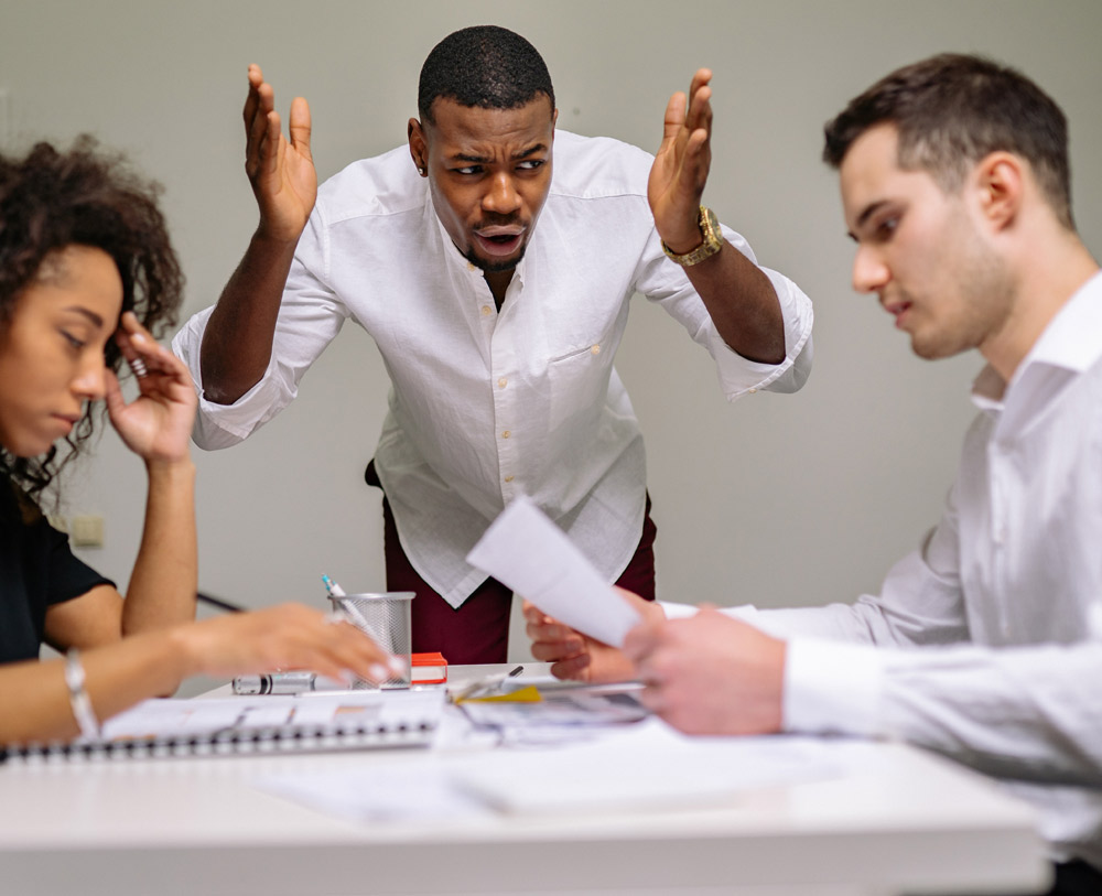 A man is seen shouting at two of his work colleagues as they are working infront of him. The two colleagues are looking at their work trying to focus as they are being distracted.