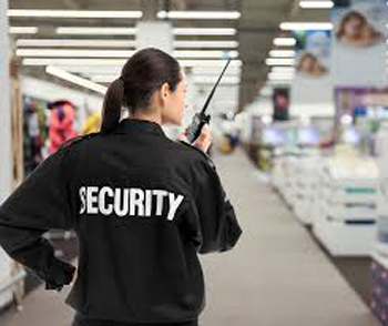 A woman seen from the back holding a walkie talkie and wearing a black jacket with 'security' on the back in white font. She is in a shop.