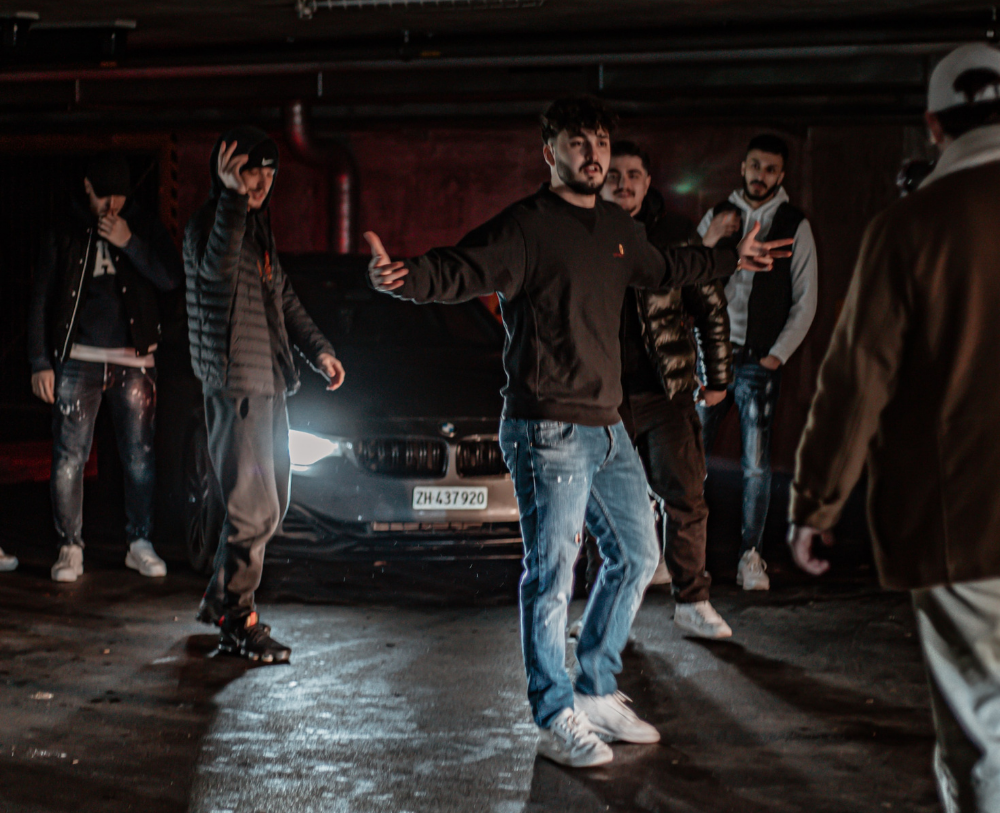 In a dark lit location, a group of young men are stood infront of a grey BMW wearing tracksuits, jeans and trainers as they approve another young man approaching them. The group have they hands in the air gesturing that there could be an altercation.